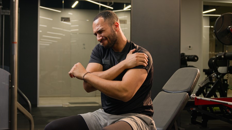 Obraz przedstawia osobę w siłowni trzymającą się za ramię, co może sugerować przeciążenie mięśni.